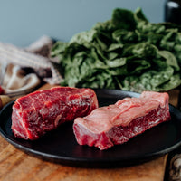Two pieces of raw Boneless Beef Short Ribs on a black plate with a variety of vegetables in the background.