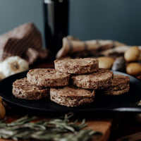 A stack of six raw Grassfed Beef Italian Breakfast Sliders on a black plate with potatoes and herbs in the background