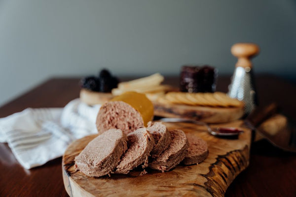 Grassfed Beef Liverwurst sliced on a wooden board with a blurred background of crackers, cheese, berries, jam, and a grader