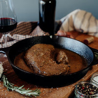 Grassfed Beef Pot Roast precooked with gravy in a cast iron skillet on a wooden cutting board with a glass of red wine and spices.