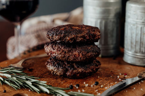 Three cooked patties of 75% Lean Ground Beef with Heart, Kidney, Liver blend in three sections on a piece of paper with a wooden surface with seasonings and rosemary in the background