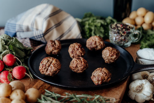 Seven Ground Lamb meatballs on a black skillet with vegetables, cloths, and herbs on a wooden surface