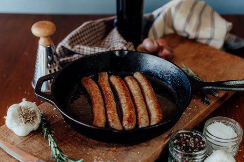 Cooked Rusti Lamb Sausage Links in a black skillet on a wooden board with seasonings and vegetables