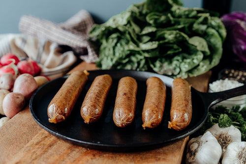 Five Sugar Free Smoked Pork Bacon Bratwurst on a black skillet with vegetables and seasonings on a wooden table