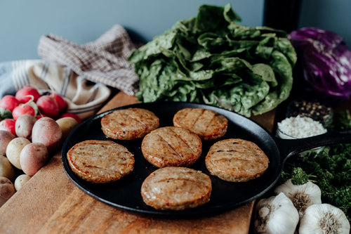 Six cooked Sugar Free Pork Breakfast Sausage Patties on a black pan with fresh vegetables in the background