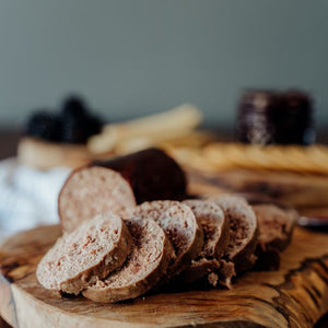 Sliced Grassfed Beef Head Cheese on a wooden cutting board with a blurred background of crackers, berries, jam, cheese, and a grader