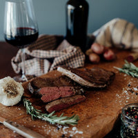 One cooked grass-fed beef liver on a cutting board and one cooked and sliced with spices, rosemary, garlic, potatoes, a grader, cloths, a bottle, and glass of wine