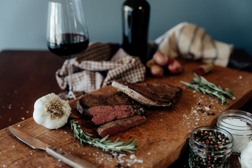 One cooked grass-fed beef liver on a cutting board and one cooked and sliced with spices, rosemary, garlic, potatoes, a grader, cloths, a bottle, and glass of wine