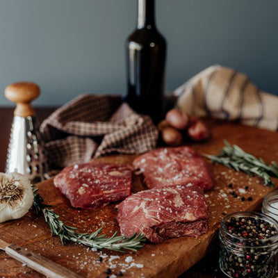 Three raw beef minute steaks on a wooden cutting board with potatoes and spices, set against a dark background.